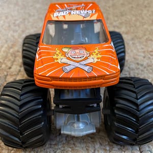 May include: Orange monster truck toy with large black tires. The truck features the text "BAD NEWS!" and a cartoon graphic of a smiling character with crossed newspapers. The toy is designed with a detailed chassis.