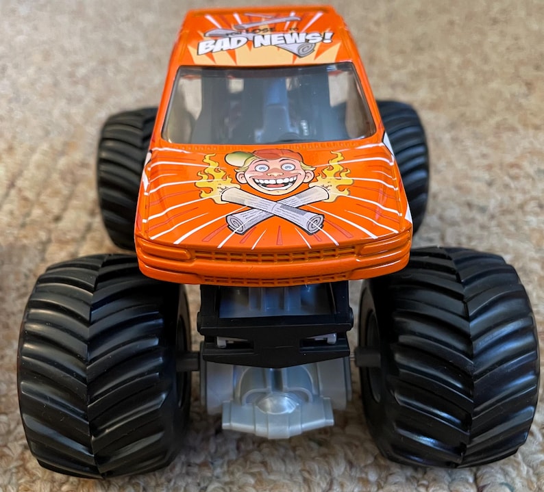 May include: Orange monster truck toy with large black tires. The truck features the text "BAD NEWS!" and a cartoon graphic of a smiling character with crossed newspapers. The toy is designed with a detailed chassis.