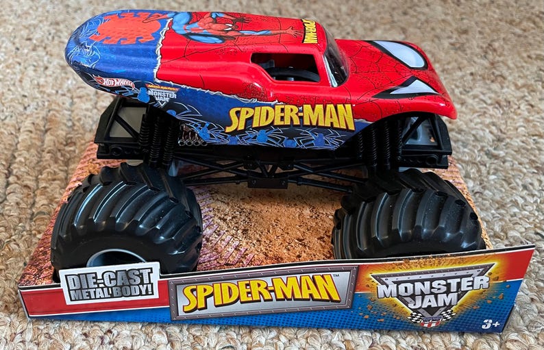 May include: A red and blue Spider-Man monster truck toy with black tires. The truck has the words "Spider-Man" and "Monster Jam" on it. The toy is on a cardboard display.