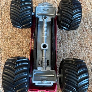 May include: A top-down view of a toy monster truck. The truck has a magenta body, a silver chassis, and four large black tires with deep treads. The tires are designed for off-road driving.
