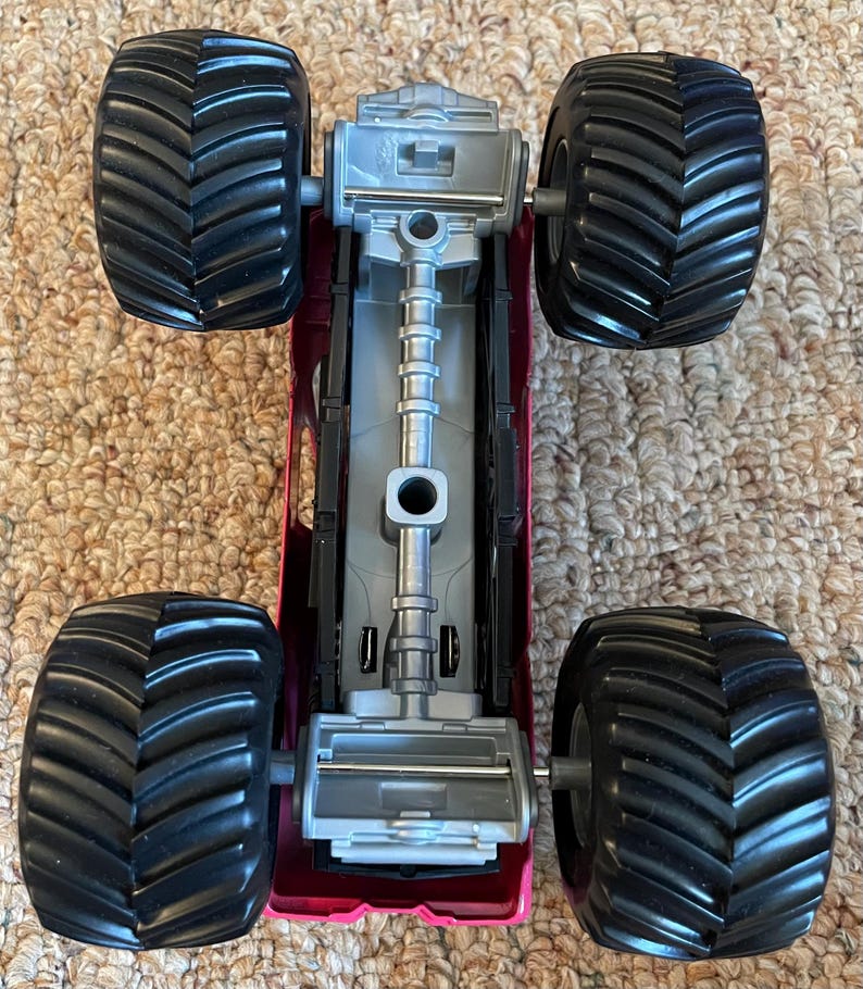 May include: A top-down view of a toy monster truck. The truck has a magenta body, a silver chassis, and four large black tires with deep treads. The tires are designed for off-road driving.