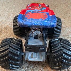 May include: A toy monster truck with a red and blue body featuring a Spider-Man graphic. The truck has large black tires and a detailed chassis. The toy is designed for play and display.