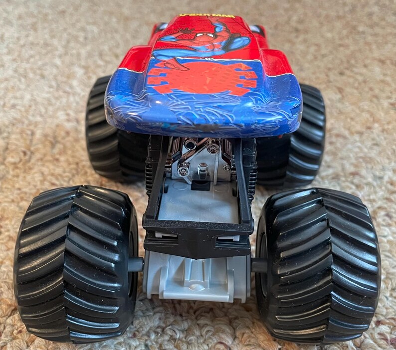 May include: A toy monster truck with a red and blue body featuring a Spider-Man graphic. The truck has large black tires and a detailed chassis. The toy is designed for play and display.