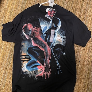 May include: Black t-shirt featuring a graphic of Spider-Man in red and blue, and a black Spider-Man. The design includes a cityscape and rain effect. The shirt has a tag attached.