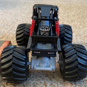 May include: A black and red toy monster truck with large, black tires. The truck has a detailed engine and suspension system. The tires are thick and have deep treads. The truck is designed to look like a real monster truck.