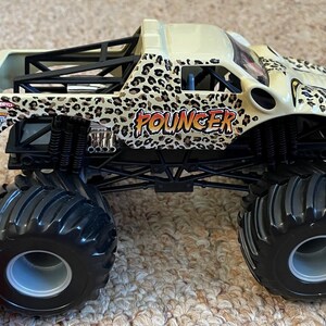 May include: A toy monster truck with a cream-colored body and black chassis. The truck features a leopard print design and the word "POUNCER" in orange. Large black tires and a roll cage complete the look.