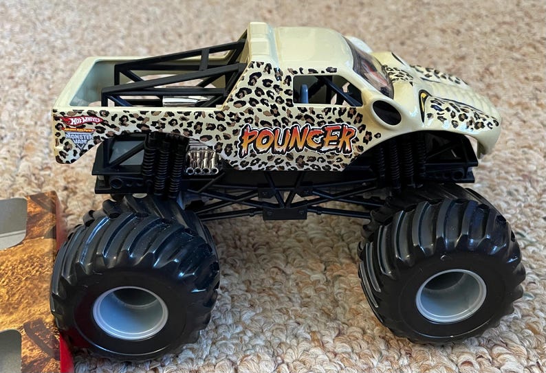 May include: A toy monster truck with a cream-colored body and black chassis. The truck features a leopard print design and the word "POUNCER" in orange. Large black tires and a roll cage complete the look.