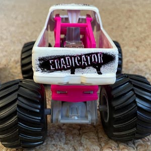 May include: A white and pink toy monster truck with large black tires. The truck has the word "ERADICATOR" in black on the back. The truck has a pink roll cage and a silver engine.