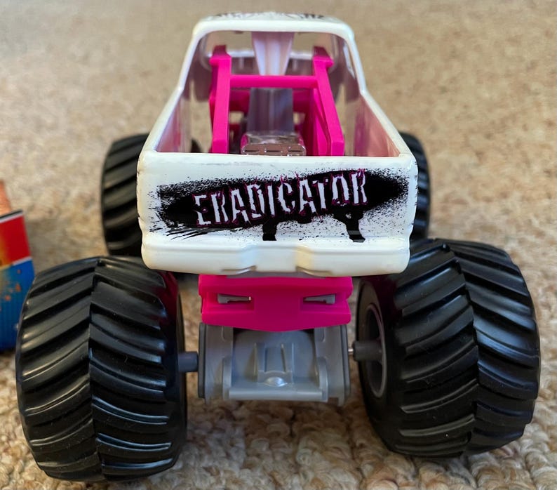 May include: A white and pink toy monster truck with large black tires. The truck has the word "ERADICATOR" in black on the back. The truck has a pink roll cage and a silver engine.