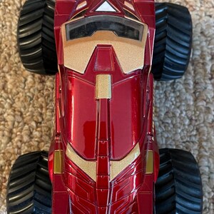 May include: A red and gold toy monster truck with large black tires. The truck has a glossy finish and features gold accents on the front and sides. The design is reminiscent of a superhero's vehicle.