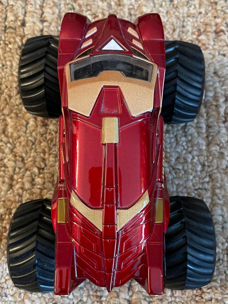 May include: A red and gold toy monster truck with large black tires. The truck has a glossy finish and features gold accents on the front and sides. The design is reminiscent of a superhero's vehicle.