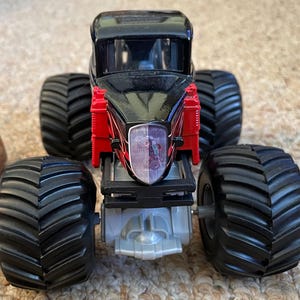 May include: A black and red monster truck toy with large, black tires. The truck has a black cab and red accents. The front of the truck features a clear headlight with a graphic inside. The tires are thick and have deep treads.