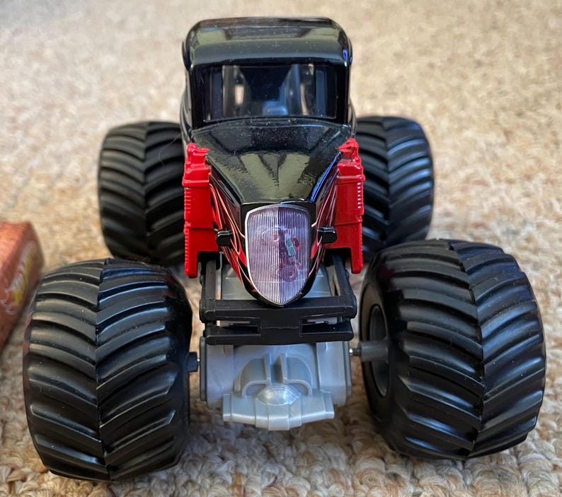 May include: A black and red monster truck toy with large, black tires. The truck has a black cab and red accents. The front of the truck features a clear headlight with a graphic inside. The tires are thick and have deep treads.