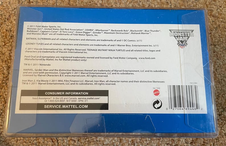 May include: A blue and white box with the Monster Jam logo. The box contains text about trademarks and consumer information. The text includes the names of Monster Jam, Batman, Superman, Looney Tunes, Teenage Mutant Ninja Turtles, and Marvel characters.