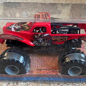 May include: A red monster truck toy with black tires and a black roll cage. The truck is decorated with pirate-themed graphics and the words "Captain's Curse". The toy is packaged in a clear plastic container.