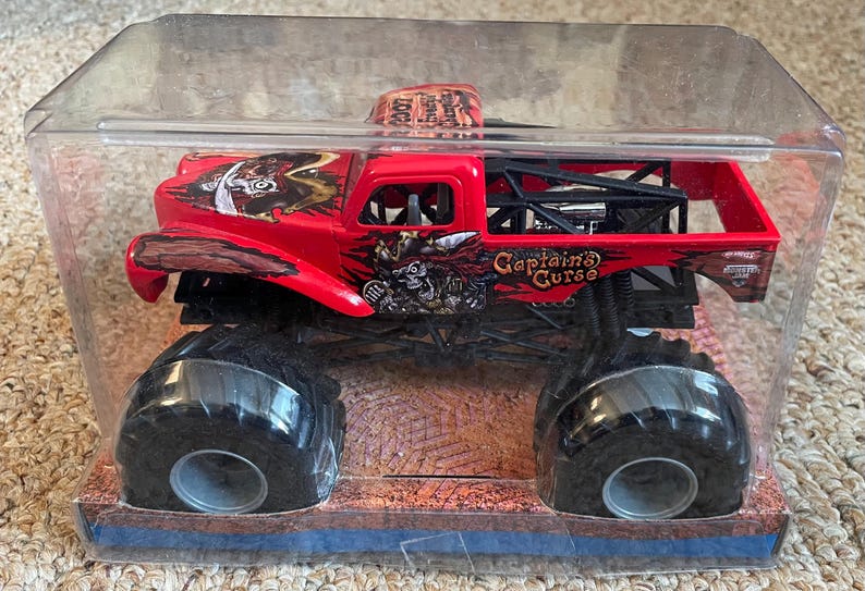 May include: A red monster truck toy with black tires and a black roll cage. The truck is decorated with pirate-themed graphics and the words "Captain's Curse". The toy is packaged in a clear plastic container.