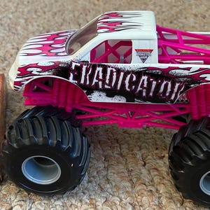 May include: A toy monster truck with large black tires and a white cab. The truck has a pink roll cage and accents, with the word "ERADICATOR" in black. The truck is decorated with a flame design in pink and white. A box with the Monster Jam logo is visible.