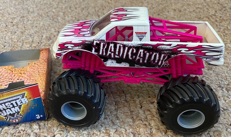 May include: A toy monster truck with large black tires and a white cab. The truck has a pink roll cage and accents, with the word "ERADICATOR" in black. The truck is decorated with a flame design in pink and white. A box with the Monster Jam logo is visible.
