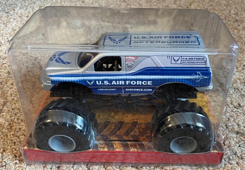 Pu&ograve; includere: Un camion mostro giocattolo della U.S. Air Force argento e blu in una confezione di plastica trasparente. Il camion presenta grandi pneumatici neri e le parole "U.S. AIR FORCE" e "AFTERBURNER" stampate sui lati.
