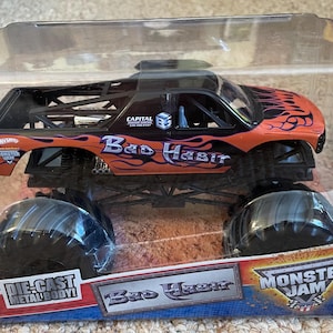 May include: Die-cast metal body monster truck toy in a clear plastic package. The truck is orange and black with the words "Bad Habit" and flame graphics. The package also features the Monster Jam logo.