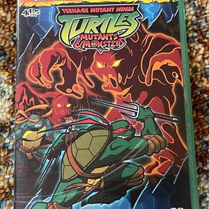 May include: A DVD case for "Teenage Mutant Ninja Turtles: Mutants & Monsters" featuring the turtles in action. The cover has a green border and displays the title with a fiery background and the text "Six Terrifying Adventures!"