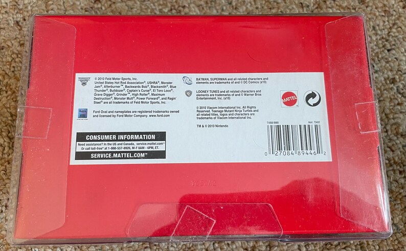 Puede incluir: Una caja roja con una cubierta de pl&aacute;stico transparente. La caja contiene texto, incluyendo "CONSUMER INFORMATION" y "SERVICE.MATTEL.COM". La caja tambi&eacute;n tiene un c&oacute;digo de barras y el logotipo de Mattel.