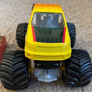 May include: A yellow and orange toy monster truck with large black tires. The truck has a black and green grid design on the roof and a red stripe. The front of the truck has a yellow grill.