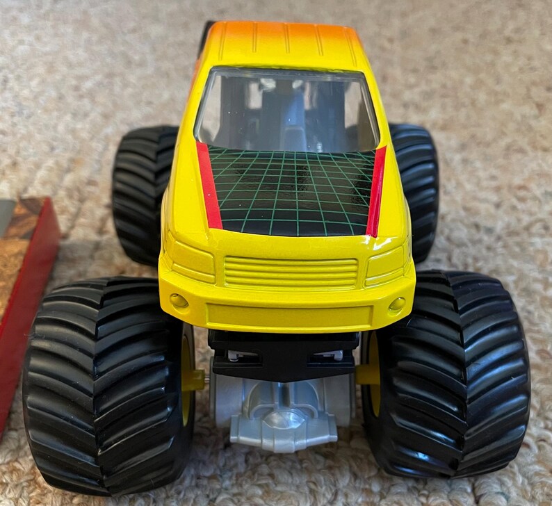 May include: A yellow and orange toy monster truck with large black tires. The truck has a black and green grid design on the roof and a red stripe. The front of the truck has a yellow grill.