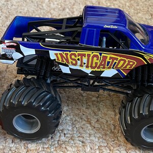 May include: A blue and black toy monster truck with the word "INSTIGATOR" in yellow and red lettering. The truck has large black tires and a black chassis. The Hot Wheels Monster Jam logo is visible on the side.