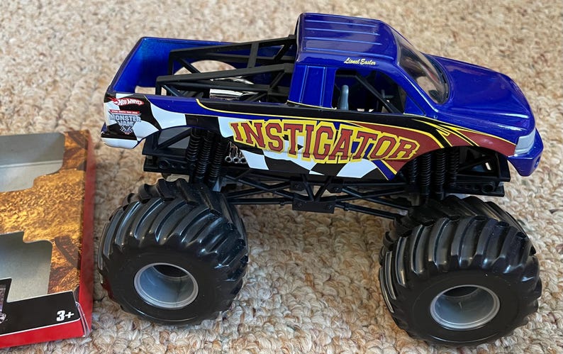 May include: A blue and black toy monster truck with the word "INSTIGATOR" in yellow and red lettering. The truck has large black tires and a black chassis. The Hot Wheels Monster Jam logo is visible on the side.