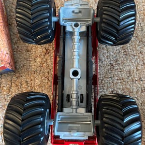 May include: A red and silver toy monster truck with four large black tires. The truck has a detailed chassis and is designed for off-road play. The tires have deep treads for traction.