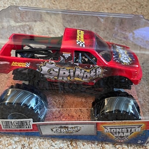 May include: A red and black die-cast metal monster truck toy, labeled "Grinder" with large black tires. The toy is in a clear plastic package with the "Monster Jam" logo.