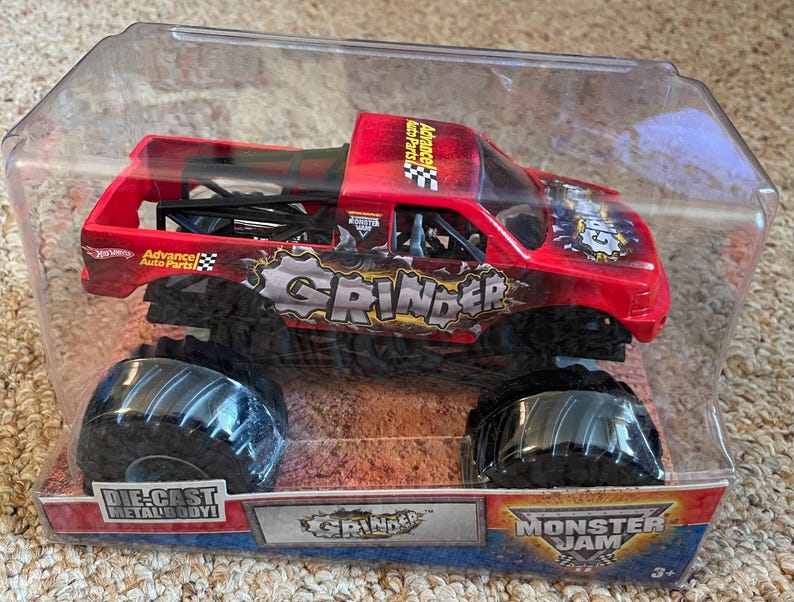 May include: A red and black die-cast metal monster truck toy, labeled "Grinder" with large black tires. The toy is in a clear plastic package with the "Monster Jam" logo.