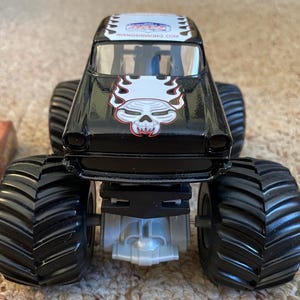 Puede incluir: Cami&oacute;n monstruo de juguete negro con neum&aacute;ticos negros grandes. El cami&oacute;n presenta un gr&aacute;fico de calavera con llamas en la parte delantera y el texto "AVENGERRACING.COM" en el techo. Es un veh&iacute;culo de juguete.