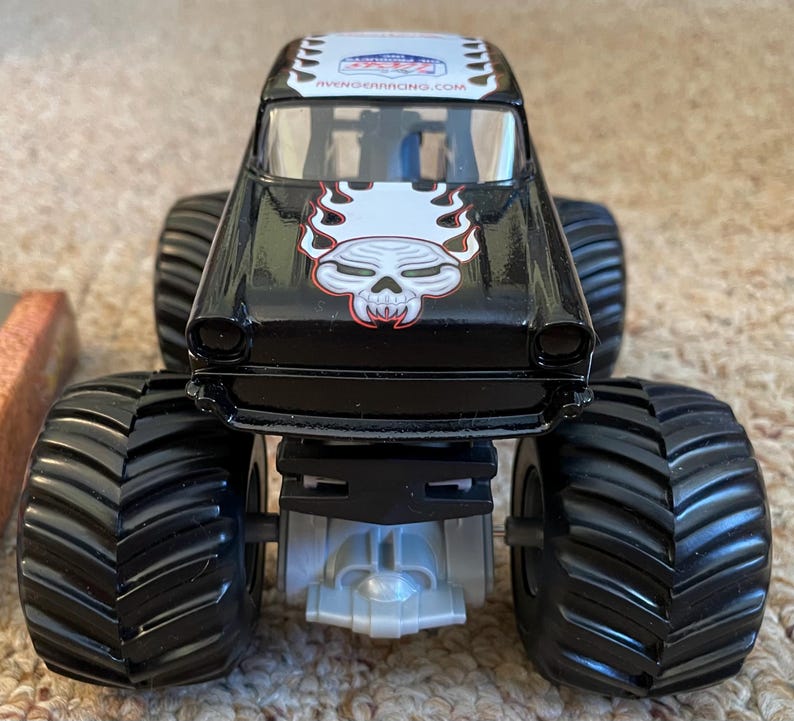 Puede incluir: Cami&oacute;n monstruo de juguete negro con neum&aacute;ticos negros grandes. El cami&oacute;n presenta un gr&aacute;fico de calavera con llamas en la parte delantera y el texto "AVENGERRACING.COM" en el techo. Es un veh&iacute;culo de juguete.