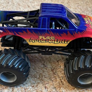Może przedstawiać: Niebiesko-czerwono-ż&oacute;łta ciężar&oacute;wka zabawka Hot Wheels Monster Jam. Posiada duże czarne opony i napis "Pure Adrenaline" z boku. Zabawka to pojazd.