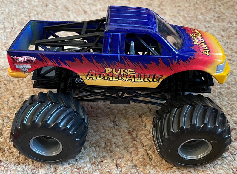 Może przedstawiać: Niebiesko-czerwono-ż&oacute;łta ciężar&oacute;wka zabawka Hot Wheels Monster Jam. Posiada duże czarne opony i napis "Pure Adrenaline" z boku. Zabawka to pojazd.