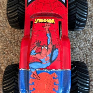 May include: A red and blue Spider-Man monster truck toy with large black tires. The truck features the Spider-Man logo and a graphic of Spider-Man. The toy is designed for play and display.