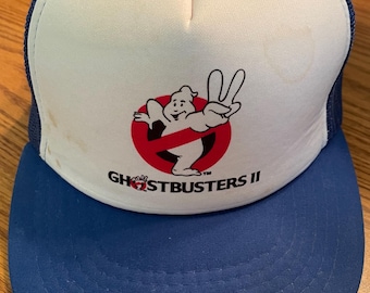 Op de afbeelding: Een blauw-witte Ghostbusters II truckerpet. Het voorpaneel is wit met het Ghostbusters-logo in rood en zwart. De klep en de mesh-achterkant zijn blauw. De pet heeft een snapback-sluiting.