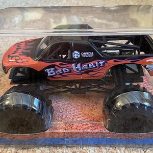 May include: A toy monster truck in a clear plastic case. The truck is black and orange with the words "Bad Habit" and flame graphics. It has large black tires and is displayed on a desert-themed base.