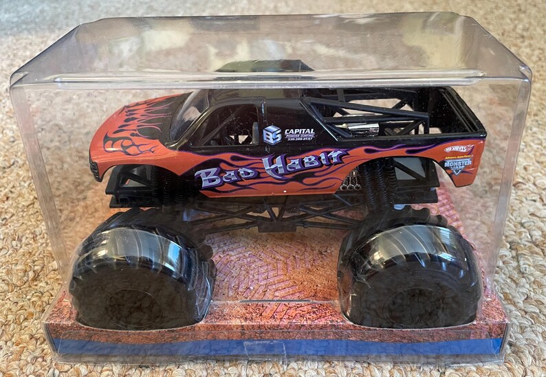 May include: A toy monster truck in a clear plastic case. The truck is black and orange with the words "Bad Habit" and flame graphics. It has large black tires and is displayed on a desert-themed base.