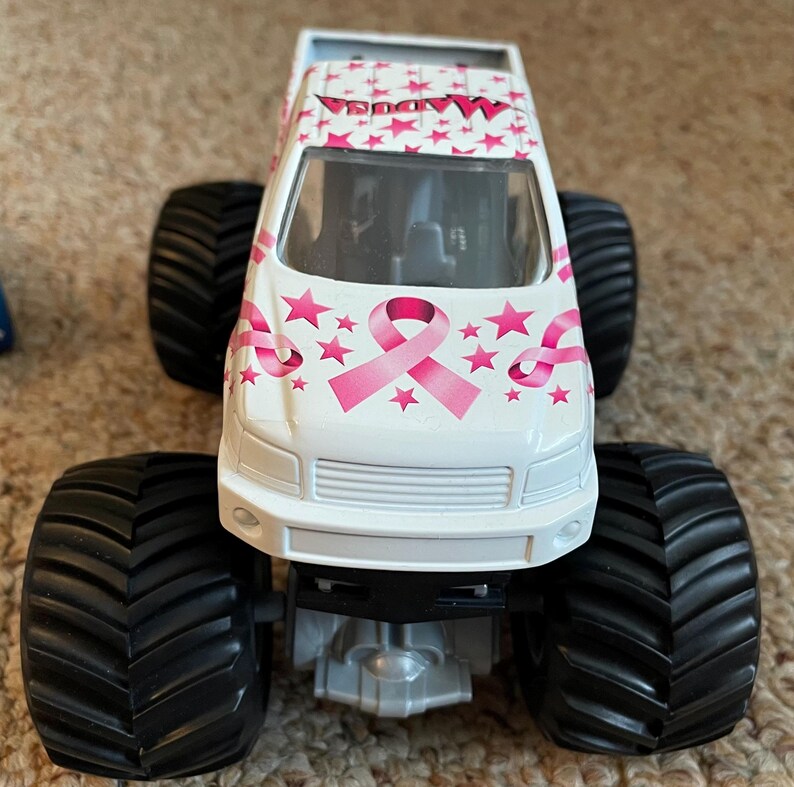 K&ouml;nnte beinhalten: Ein wei&szlig;er Spielzeug-Monstertruck mit gro&szlig;en schwarzen Reifen. Der Truck hat rosa Sterne und ein rosa Schleifenmuster, mit dem Wort "MADUSA" auf dem Dach. Der Truck ist ein Spielzeug.