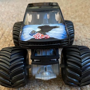 May include: Black monster truck toy with large black tires. The hood features a graphic of an eagle, the American flag, and a blue sky background. The truck has a silver chassis.