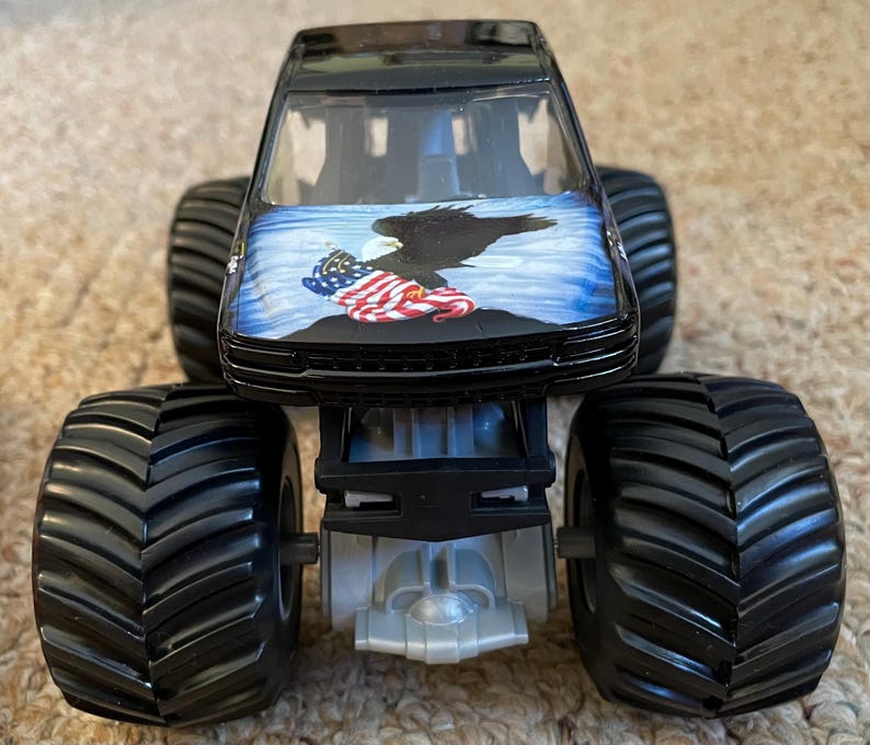 May include: Black monster truck toy with large black tires. The hood features a graphic of an eagle, the American flag, and a blue sky background. The truck has a silver chassis.