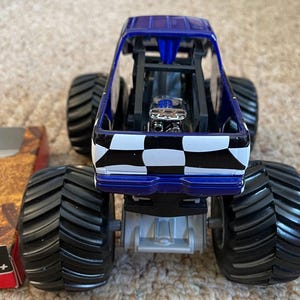 May include: A blue and black monster truck toy with large black tires. The truck features a checkered pattern on the back and a detailed engine. The toy is designed for children aged 3 and up.