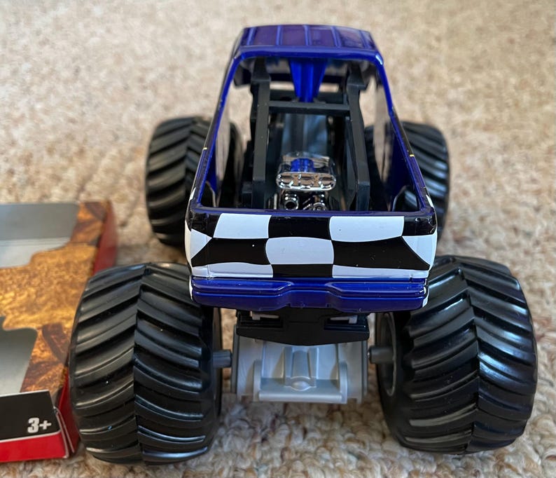 May include: A blue and black monster truck toy with large black tires. The truck features a checkered pattern on the back and a detailed engine. The toy is designed for children aged 3 and up.