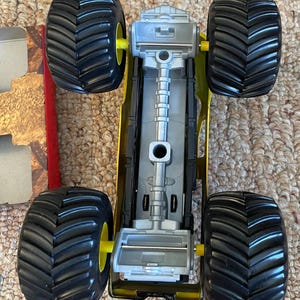 May include: A yellow and black toy monster truck with large black tires and yellow wheel hubs. The truck has a silver chassis and is viewed from above, showcasing its detailed design. The tires have a deep tread pattern.