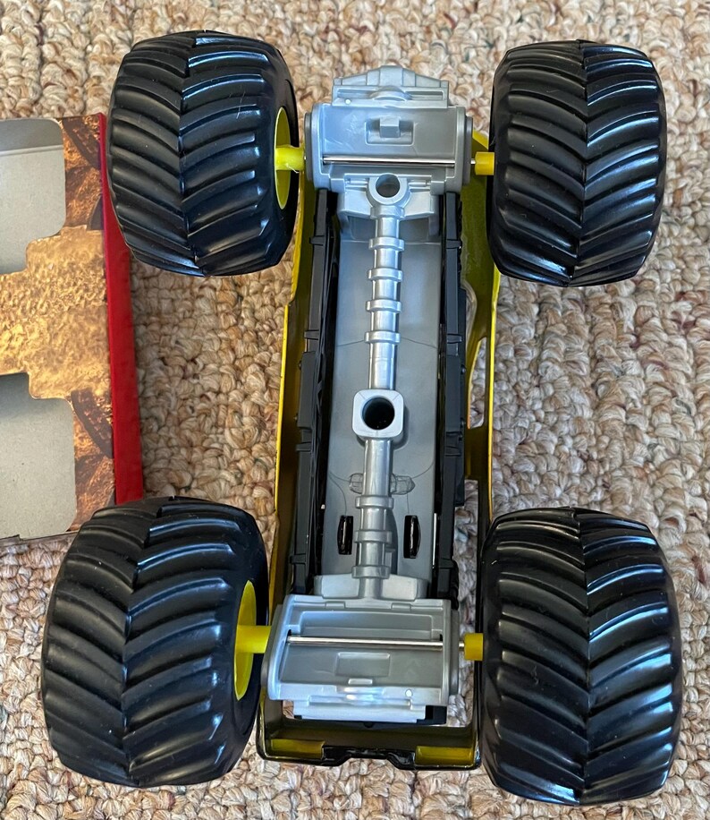 May include: A yellow and black toy monster truck with large black tires and yellow wheel hubs. The truck has a silver chassis and is viewed from above, showcasing its detailed design. The tires have a deep tread pattern.