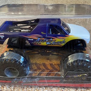 May include: A Hot Wheels monster truck toy in a clear plastic package. The truck is primarily dark blue with a white cab and yellow and orange flame graphics. The truck is labeled "Air Shock" and has large black tires.