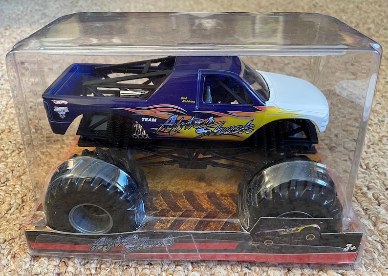May include: A Hot Wheels monster truck toy in a clear plastic package. The truck is primarily dark blue with a white cab and yellow and orange flame graphics. The truck is labeled "Air Shock" and has large black tires.